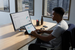 Adult male in an office setting sitting at a desk with his hand poised over the keyboard of a black Microsoft Surface Pro 7 in laptop mode. Microsoft Excel visible on labptop and Microsoft PowerBI screen seen on monitor.