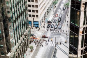 Downtown city street view from high angle inside office building. Large groups of pedestrians walk across street at crosswalk as car traffic flows through stoplight at busy intersection.