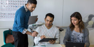 Education Color Photo of male educator using device with 6th-9th classroom of male and female students at desks with devices.