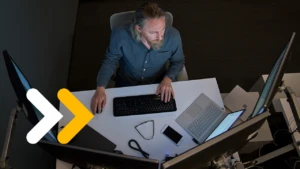 Top-down view of a bearded man in a gray/blue shirt seated at a desk working on a Surface laptop connected to three large monitors.