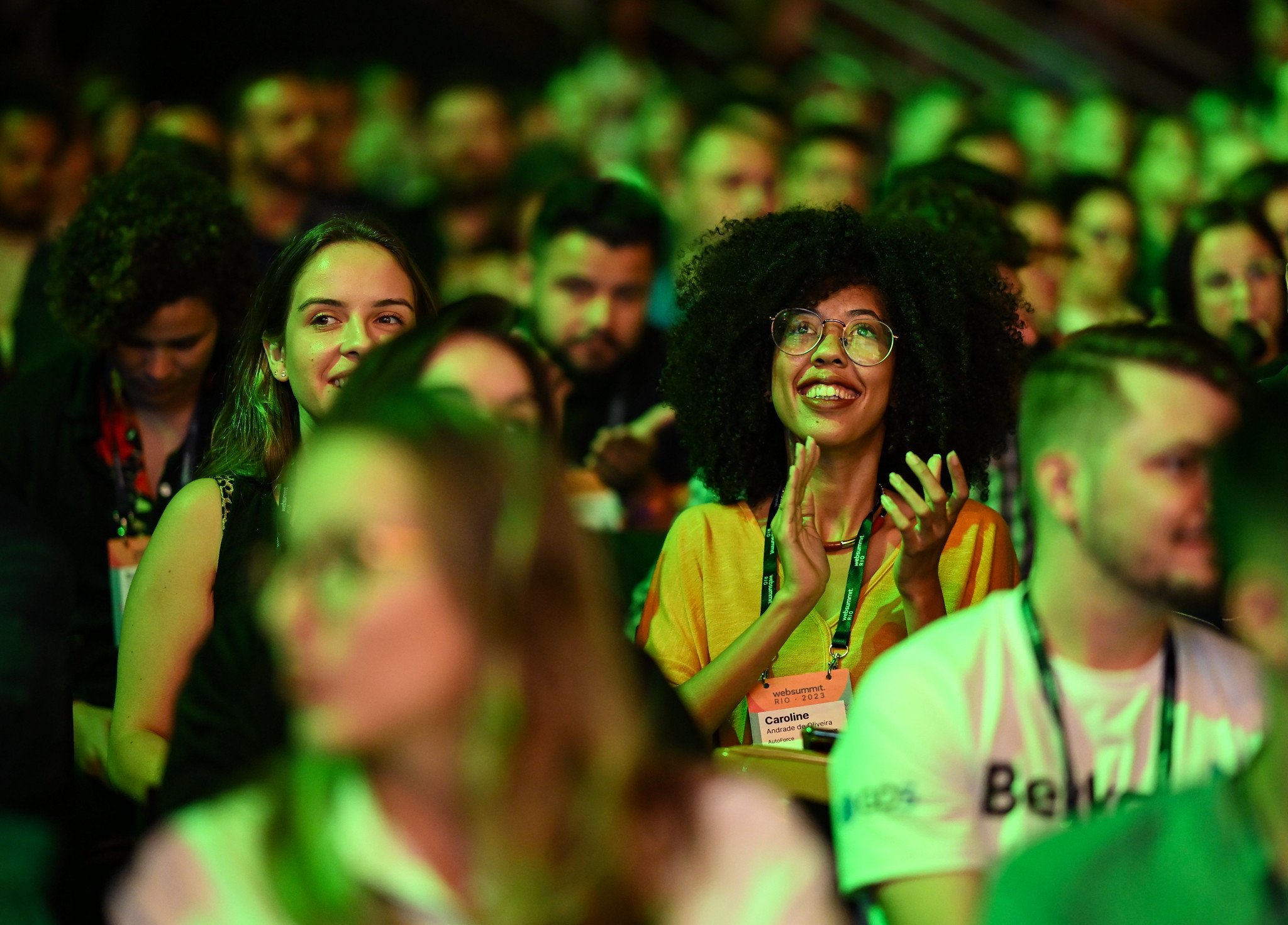 A crowd of seated people. The focus is on one person who is smiling, looking upwards – seemingly at a stage – and clapping.