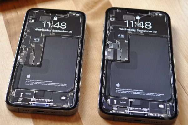 An iPhone 13 Pro and an iPhone 13 Pro Max with teardown wallpapers applied to their lock screens resting on a light-colored wood background.