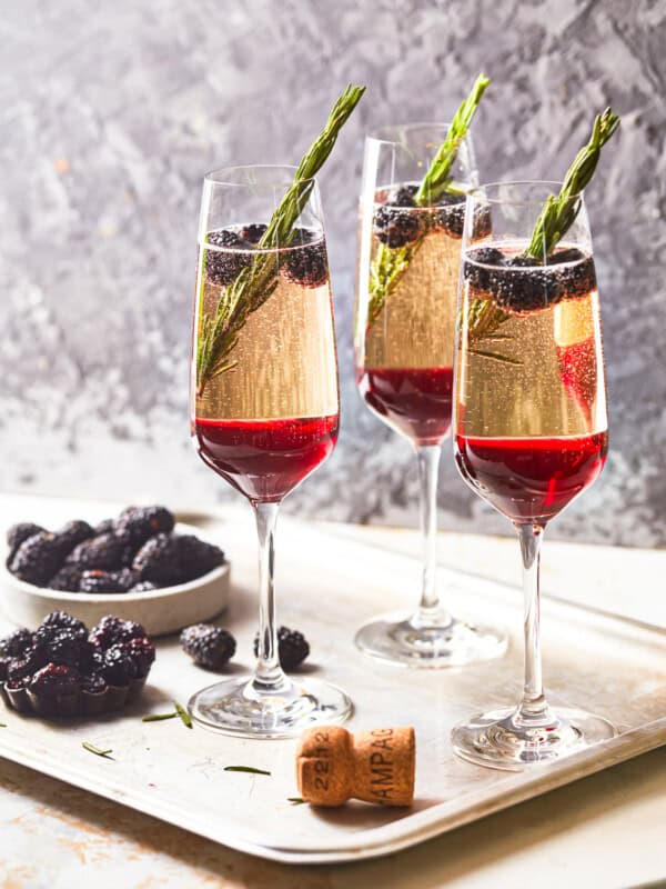 side view of 3 blackberry champagne cocktails on a metal tray with a bowl of blackberries and a champagne cork.