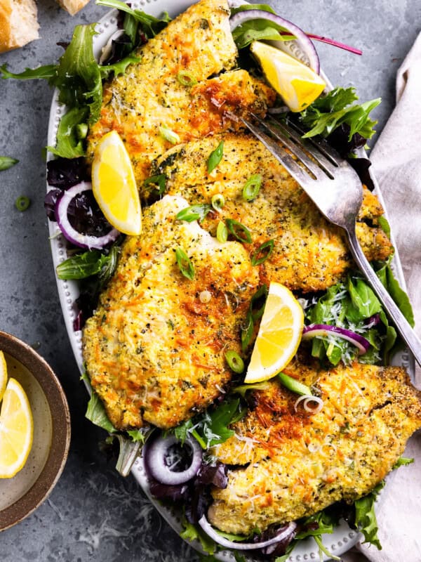 overhead view of parmesan crusted tilapia filets on an oval platter with salad greens, lemon wedges, and a fork.