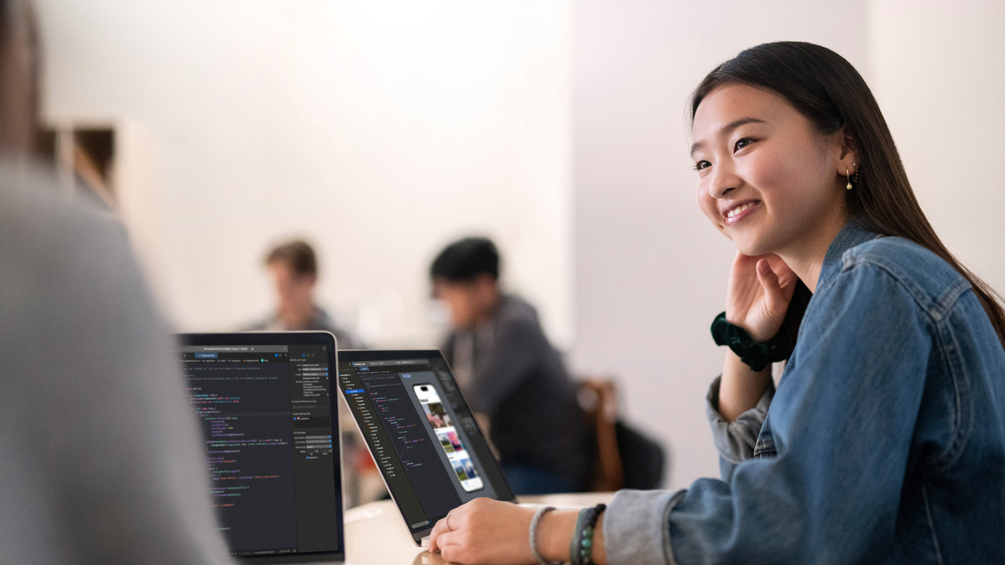 A developer wearing a denim jacket sits in front of an open laptop at a table in a room with white walls. They smile at the person next to them, who is off camera. Other developers sit in the background.