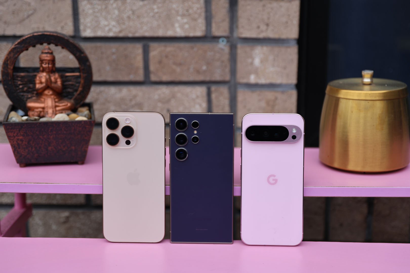 Shot of the iPhone 16 Pro Max, Samsung Galaxy S24 Ultra, and Google Pixel 9 Pro on a pink desk with a Buddha water ornament and a brass jar