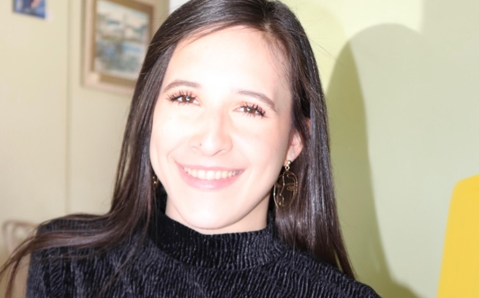 Photo of Sarai smiling to the camera. She's wearing a black blouse and big gold earrings in the shape of abstract faces.