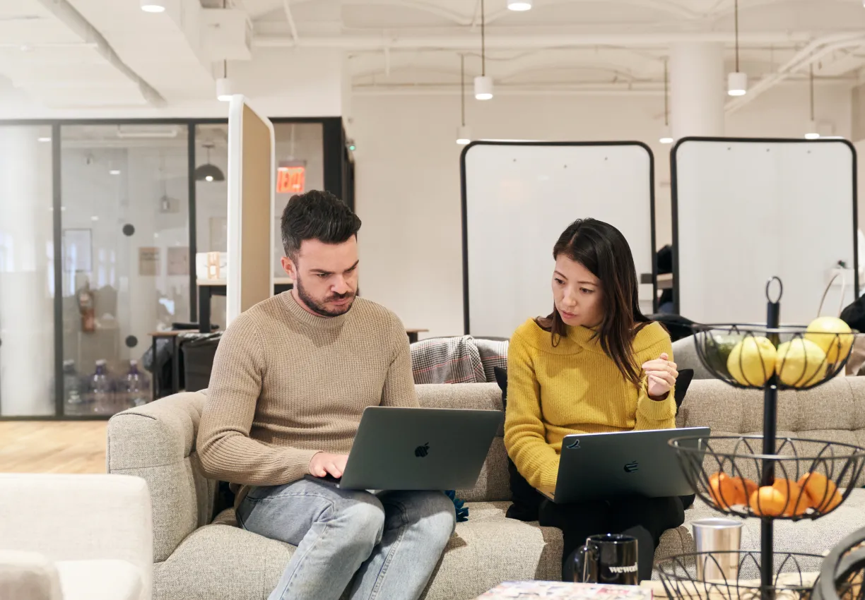 2 people working on their laptops next to each other, seated on a couch. They seem to be working through something together. 