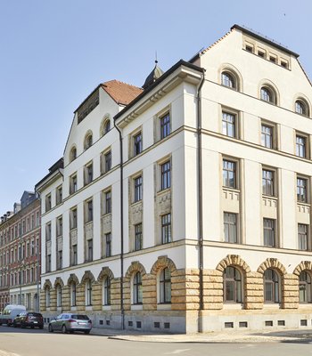 Sommerliche Aufnahme eines fünfstöckigen Gründerzeitgebäudes vor strahlend blauem Himmel. Das Gebäude ist ein Eckhaus und wird von zwei Straßen flankiert. Linker- und rechterhand stehen große grüne Laubbäume.