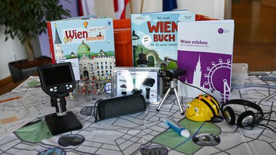 Inhalte der verschiedenen Digiboxen sind auf einem Tisch aufgestellt: Wien-Matte, Bee-Bot, Audioaufnahmegerät, Bücher, ein digitales Mikroskop, ein "Wien erleben"-Booklet und einiges mehr.