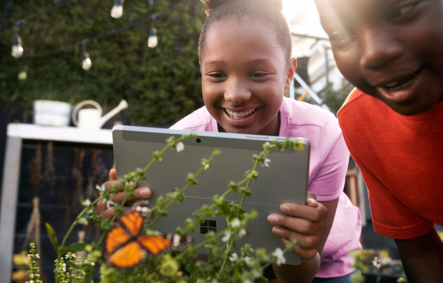 Female K-12 student in a garden holding a Surface Go while taking a picture of a butterfly. Smiling male K-12 student stands beside her.