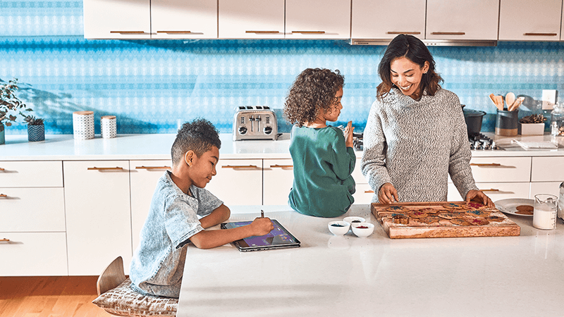 Seorang ibu berdiri dan dua anak duduk bersama di dapur.