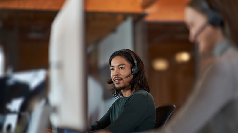 Pria dengan rambut panjang mengenakan headset, bekerja di depan komputer di sebuah kantor, dengan seorang rekan kerja di latar belakang