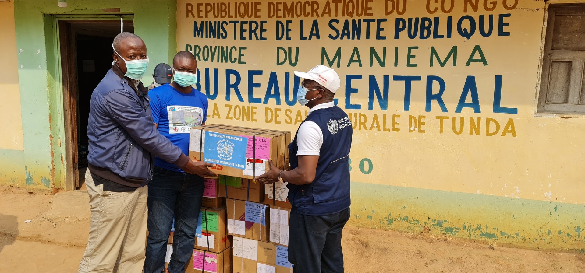 Three men wearing medical masks pose with boxes of medical supplies