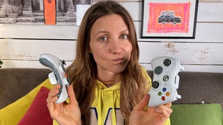 Reporterin Jana sitzt auf einer Couch. Sie hält zwei Controller in den Händen und schaut fragend in die Kamera.