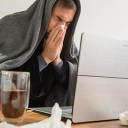 Erkälteter Mann mit Decke über dem Kopf im Homeoffice.