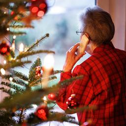 Symbolbild: Eine ältere Dame steht mit einem Telefon am Ohr neben einem geschmückten Weihnachtsbaum und blickt aus einem Fenster der Wohnung (Quelle: dpa / Matthias Balk).