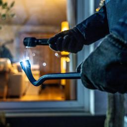 Ein Einbrecher späht von der Gartenseite her eine Wohnung aus, hält Werkzeug und Taschenlampe in der Hand.