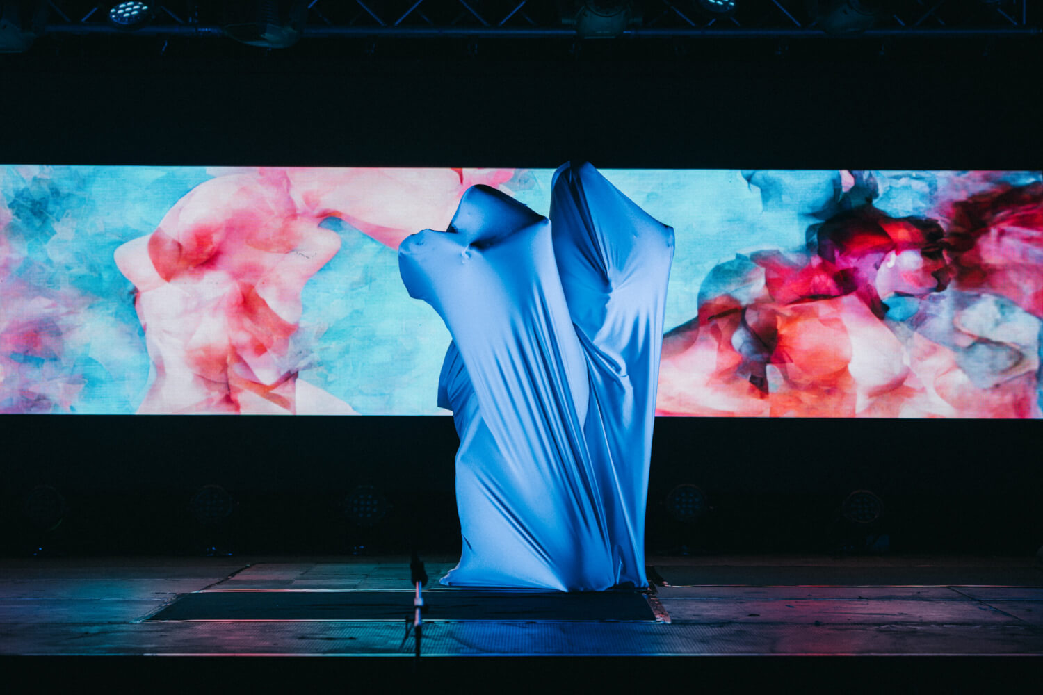 Drei Figuren unter einem Tuch silhouettenhaft zu erkennen, dahinter ein rot-blauer Screenhintergrund