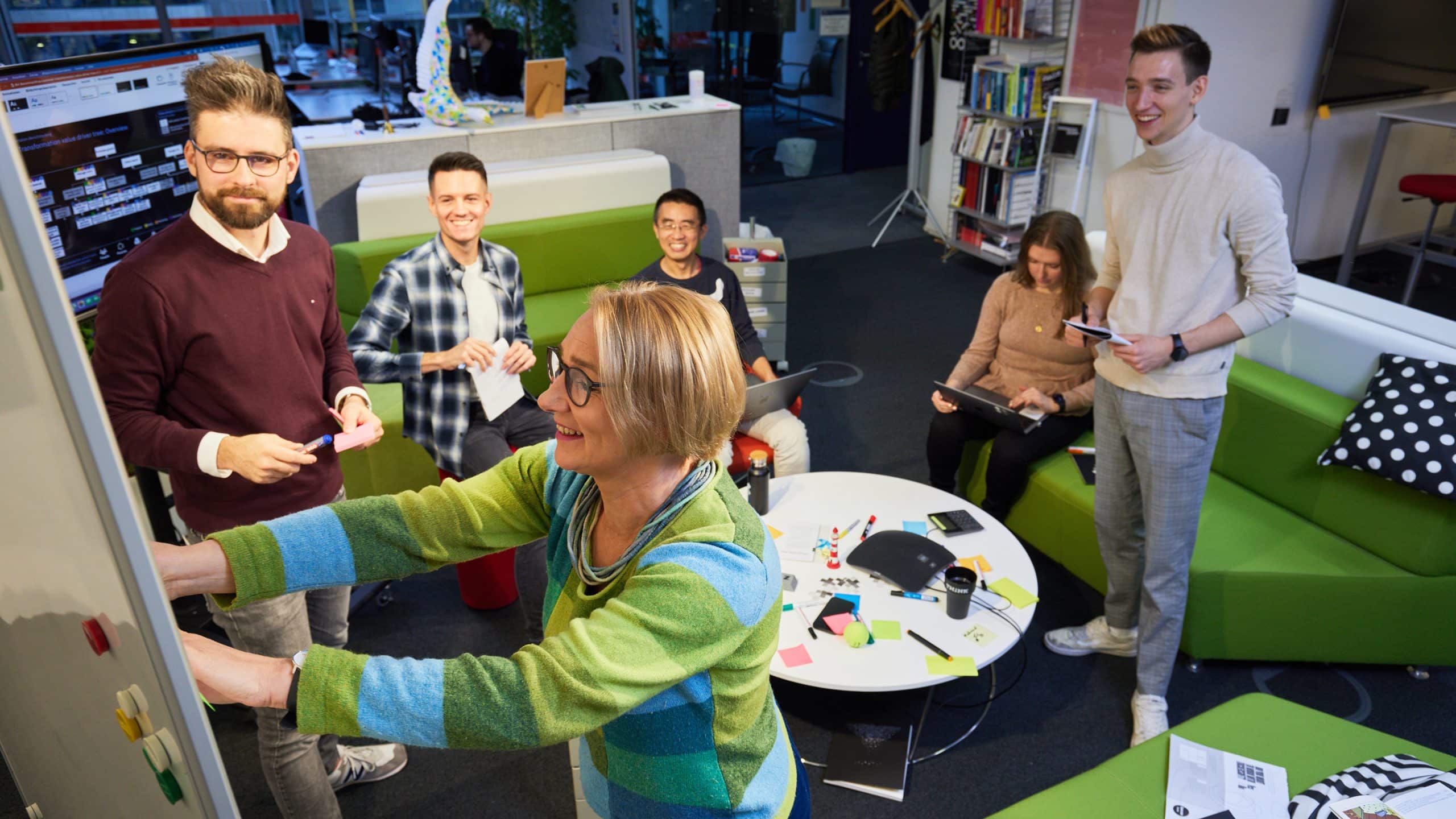 Vier Menschen arbeiten in der Gruppe. Drei sitzen und eine Frau steht an der Tafel und klebt ein Post-It ran.