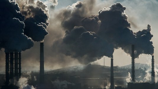 Das Foto zeigt insgesamt acht Schornsteine, aus denen viel Dampf austritt. Der Rauch zieht nach oben und etwas nach links. Im Hintergrund ist ein Hügel, davor befinden sich noch weitere Schornsteine.