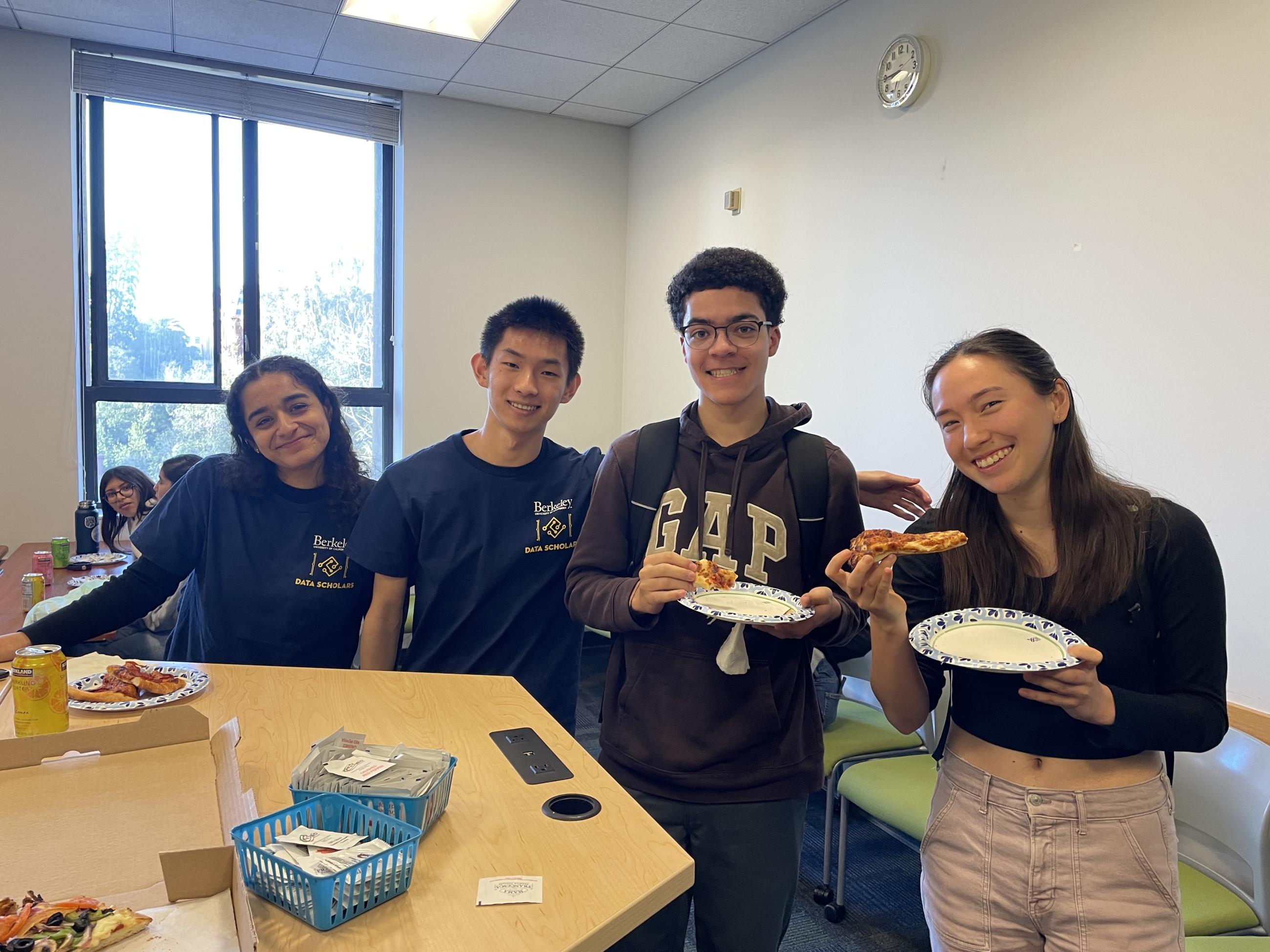 Four Data Scholars students, some holding paper plates with pizza slices, lean in toward each other and smile at the camera