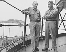 Black and white photograph of two men, one young and one old, both in khakis standing near a railing on some kind of elevated structure