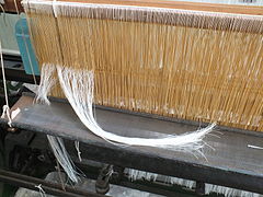 Very similar healds, with the wooden staves threaded through them top and bottom, and the warp threads in the process of being drawn in (that is, threaded through the eyes of the healds)