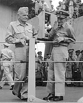 Black and white photograph of two men in khakis standing near a ship's railings, with many men a little behind them on the deck