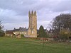 Yellow stone church tower above other buildings of the same stone. In the foreground is a grassy field with cows.