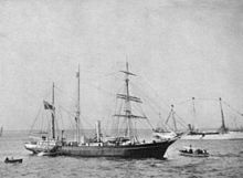 A three-masted ship with sails furled, short funnel amidships, flag flying from the stern on left of picture. Two small boats are close by, and a larger vessel decked with bunting is visible in the background.