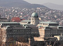 Entourant un dôme, cinq grands bâtiments à la façade rose et aux fenêtres et baies symétriques cernées de blanc se nichent derrière un village et au pied d'une montagne