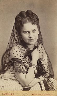 Photograph of a young woman with pinned up hair wearing a white dress and a partially transparent scarf