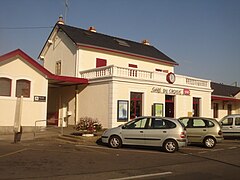 Vue d’une petite gare aux murs blancs, trois voitures garées devant.