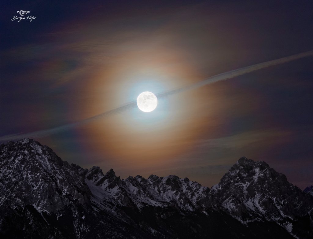 Foto des Vollmondaufgangs über schneebedeckten Gipfeln am Horizont. Der Himmel ist von einer dünnen Wolkendecke überzogen, durch den der Mond leuchtet. Er ist von einem farbigen Hof (innen bläulich und außen rotbräunlich) umgeben. Ein Kondenstreifen eines Flugzeugs verläuft von links unten nach rechts oben quer über den Mond. Ein Klick auf das Bild lädt die höchstaufgelöste verfügbare Version.