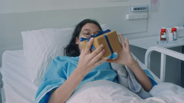 Medium shot of excited Caucasian girl in glasses and with nasal cannula receiving present from brown teddy bear while faceless parent visiting her on recovery period in childrens clinic