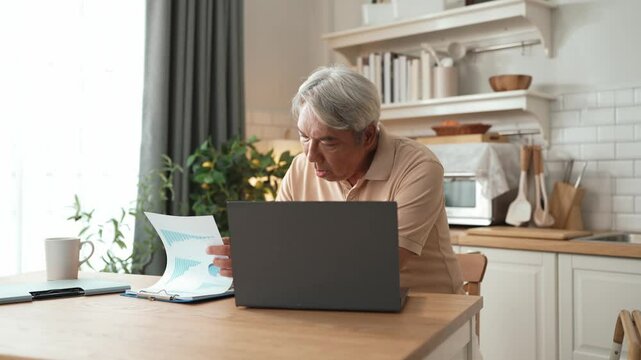 Senior business man working on laptop while checking at document at home. Middle aged manager writing report and looking at financial chart by using computer while sitting at living room. Myrmidon.