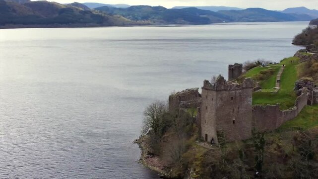 Drone footage capturing the iconic Urquhart Castle on the shores of Loch Ness in Scotland. The medieval ruins sit against a backdrop of calm waters and rolling hills, showcasing the scenic Highlands.