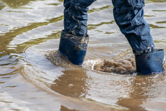 Kind mit blauer Hose und blauen Gummistiefeln watet nach einer Überschwemmung wegen Deichbruch durch das schlammige Hochwasser