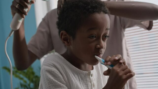 African American little boy brushing his teeth while his mom drying his hair with hair dryer, getting ready for school or kindergarten in bathroom early in morning