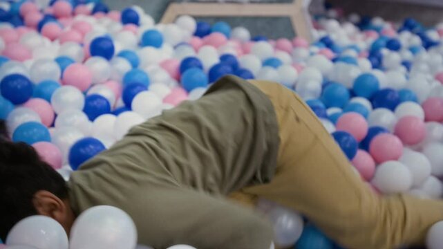 Handheld shot of small black boy enjoying playful moment in ball pit on indoor playground