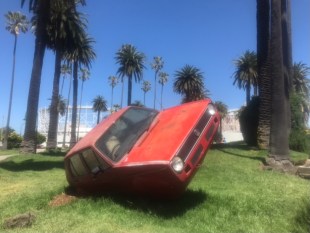 A red car installation that is perched on one side on grass.