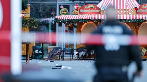 Das Bild zeigt einen leeren Stand auf dem Weihnachtsmarkt in Halle am Tag nach dem Anschlag. Im Vordergrund ist unscharf ein rot-weißes Absperrband mit der Aufschrift "Polizei" abgebildet, davor ebenfalls unscharf ein Polizist in blauer Uniform zu sehen.