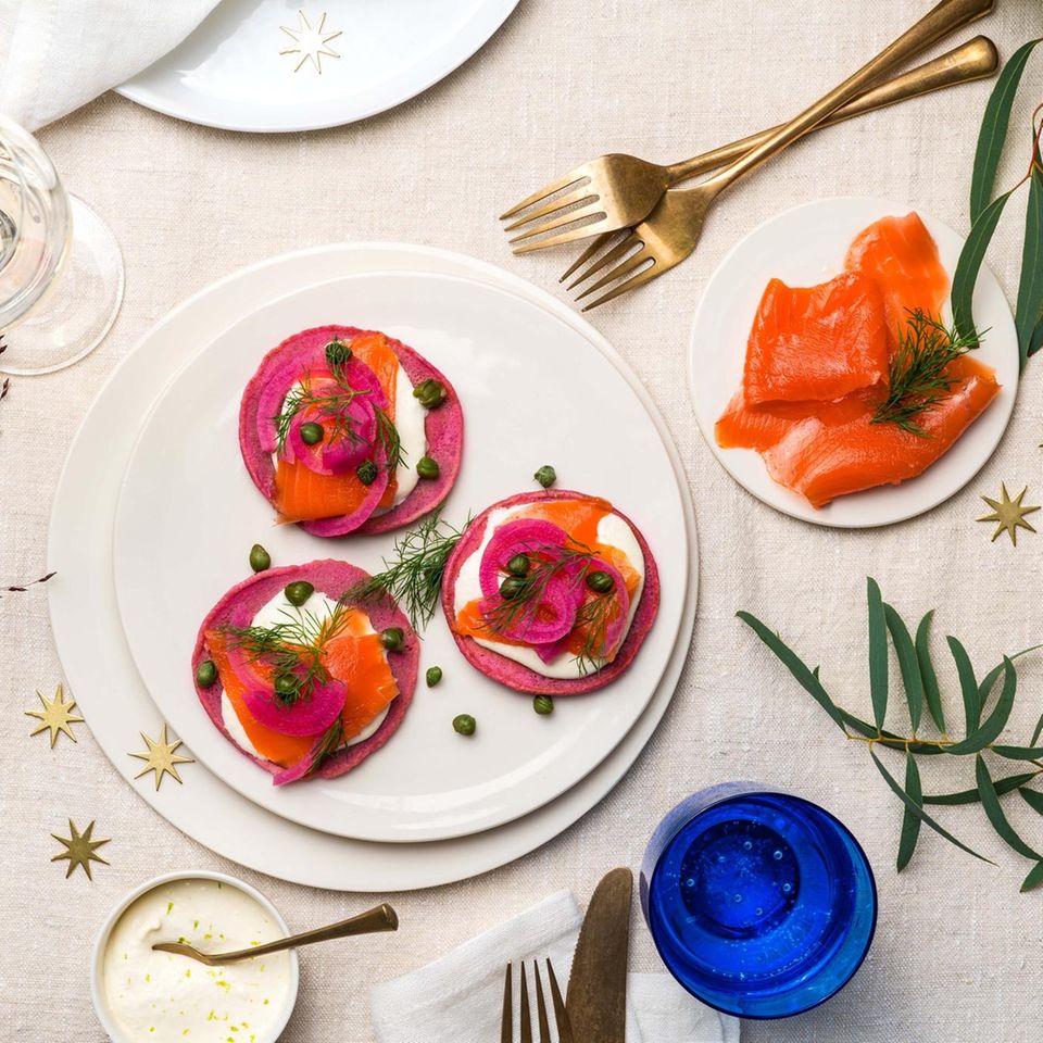 Auf einem weißen Teller sind 3 pinke Blinis mit Lachs und der Creme angerichtet, daneben steht ein Teller mit Lachs