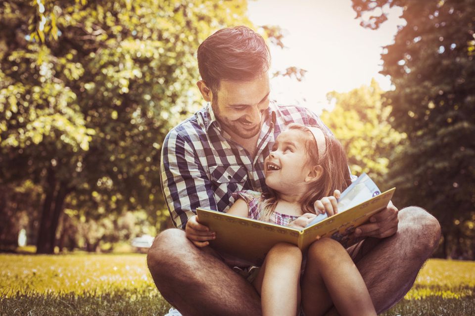 Kinderbücher für den Urlaub: Vater und Tochter lesen Buch im sommerlichen Park.