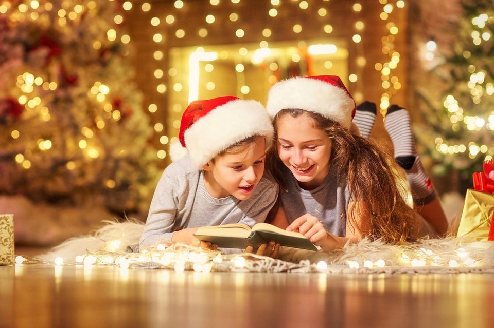 Zwei Kinder liegen mit Weihnachtsmützen an Weihnachten auf dem Teppich und lesen freudig ein Buch.