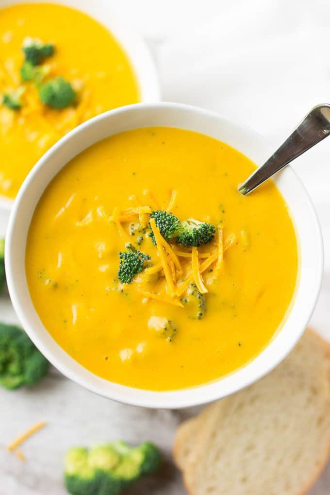 two bowls of Broccoli Cheese Soup on a white counter top with two slices of bread on the side