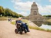 Eine Frau fährt im Rollstuhl den Weg vor dem Völkerschlachtdenkmal entlang, welches man im Hintergrund sieht.