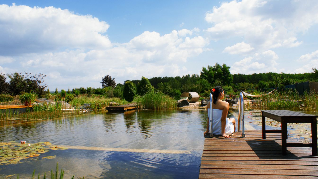 Das Bild gibt den Blick frei auf einen Seerosenteich und eine auf einem Steg sitzende Frau in einem Saunahandtuch