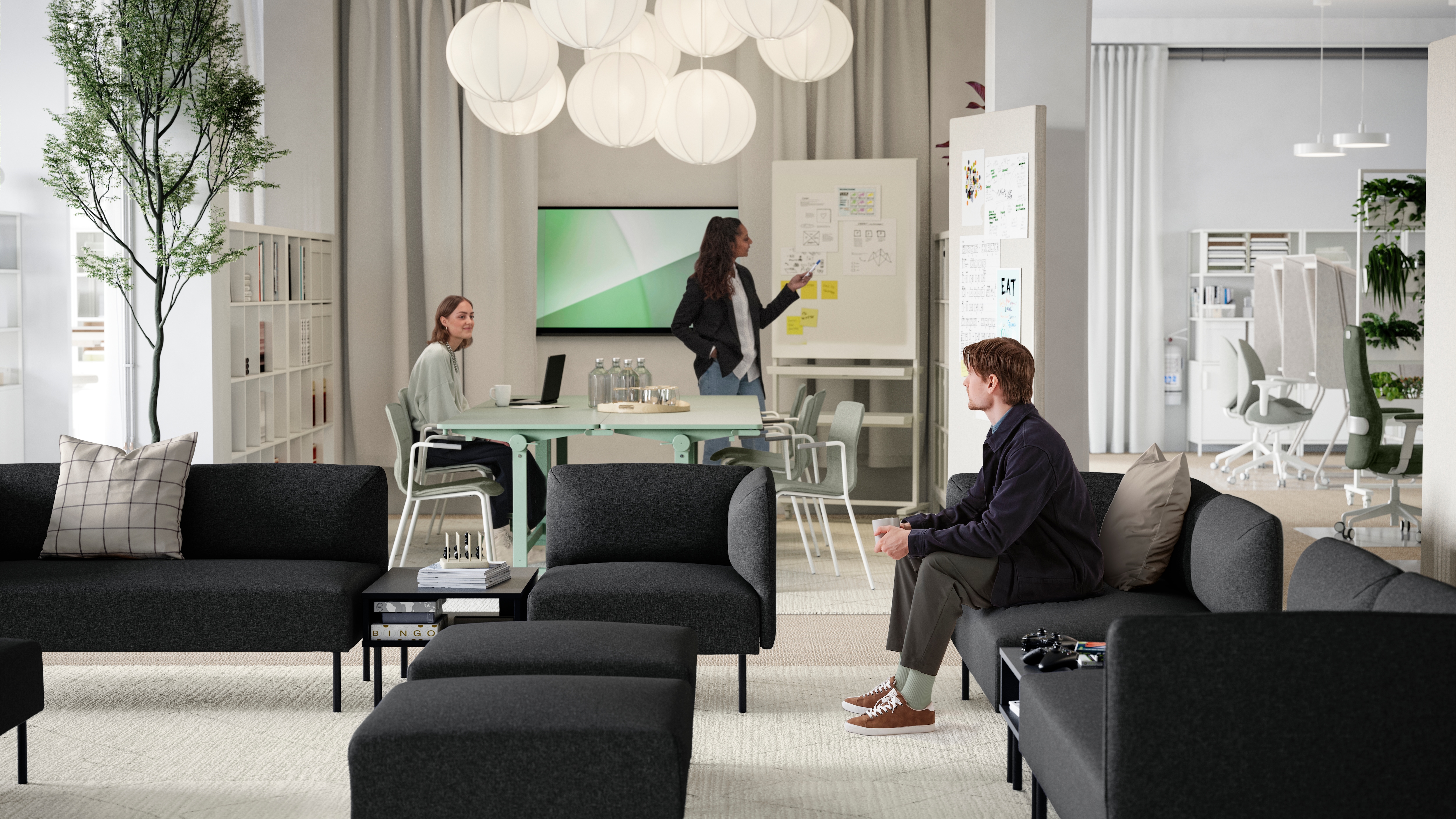 A young man sitting in a lounge area of several dark grey LILLEHEM sofa modules, TUNSTA tables and a meeting area behind it.